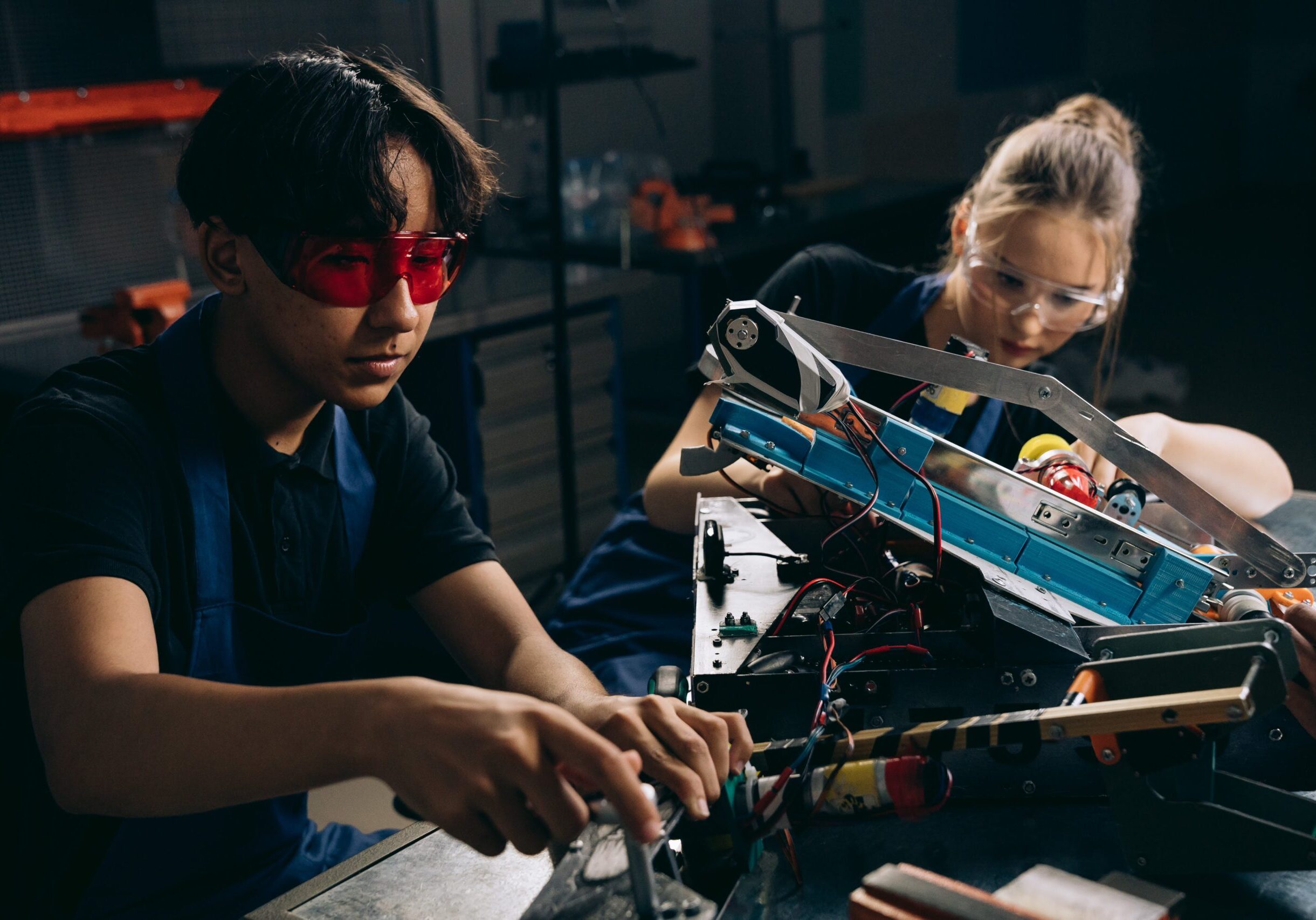 Students wearing safety gear work on robotics project, with one student in red glasses examining mechanical components