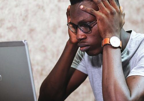 Frustrated student with glasses holding head in hands while looking at laptop screen