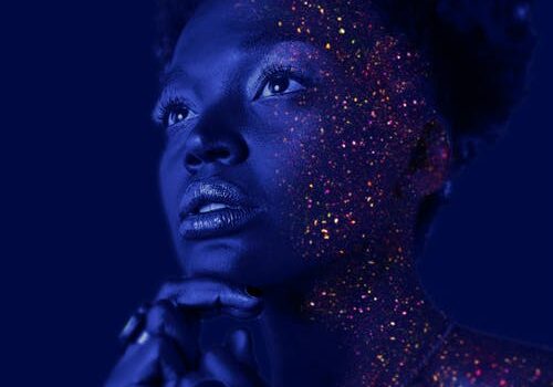 Portrait bathed in blue light with glitter effects on skin, looking upward thoughtfully with hand under chin.