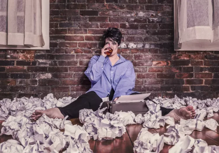 Writer in purple shirt sits against brick wall surrounded by crumpled paper, drinking from glass while working on typewriter.