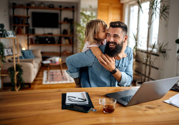 Father working from home shares joyful moment with young daughter who hugs him at wooden desk with laptop.