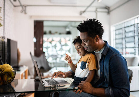 Father and young son smile while looking at laptop screen together in bright modern home office setting