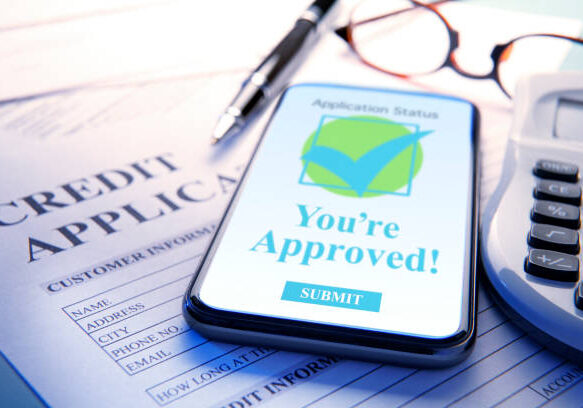 Smartphone displaying 'You're Approved!' message on credit application form with pen, glasses, and calculator