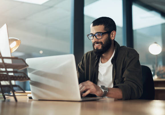 Professional in glasses smiling while working on laptop in modern office space with natural lighting