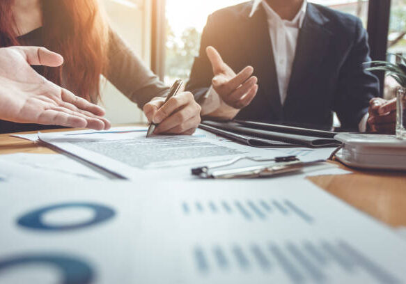 Business professionals reviewing and signing documents with financial charts and graphs on a desk in sunlit office setting