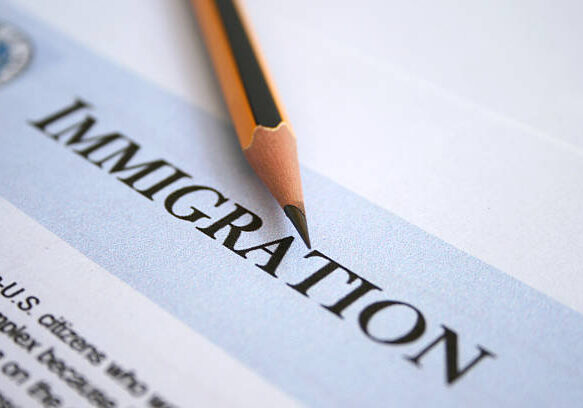 A sharp pencil point rests on a document, positioned above the word 'IMMIGRATION' printed in capital letters