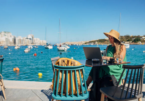 Digital nomad in green dress and sun hat works on laptop at waterfront café overlooking Mediterranean marina with boats"