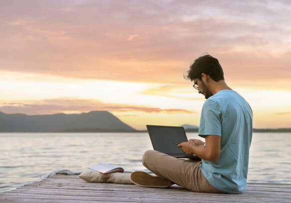 Person working on laptop while sitting on wooden dock at sunset with mountains and water in background