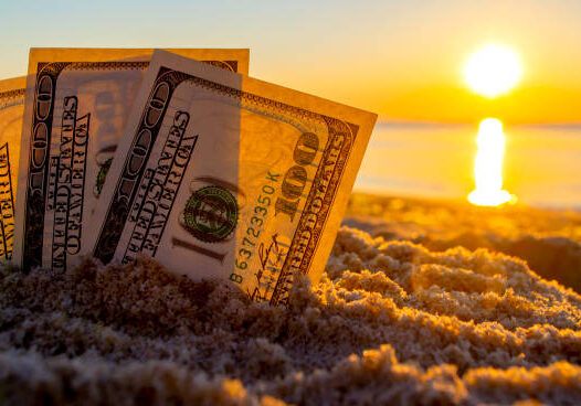 US dollar bills planted in beach sand against an orange sunset over water