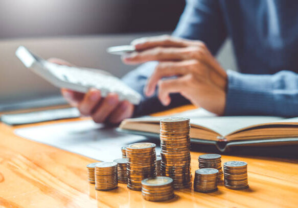 Person managing finances with stacks of coins on wooden desk while using smartphone and reviewing documents