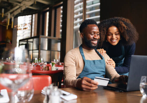 Couple smiling while looking at laptop and credit card in restaurant, man wearing blue apron and beige sweater