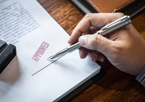 Hand holding silver pen signing document with red approval stamp on wooden desk next to rubber stamp