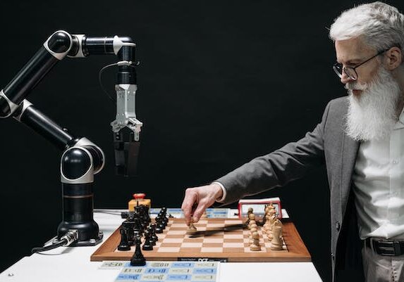 A person with a white beard plays chess against a robotic arm on a chess board in a dramatic black and white setting.