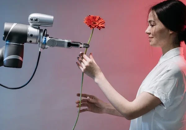 Woman in white shirt offers red gerbera daisy to robotic arm against gradient blue and pink background