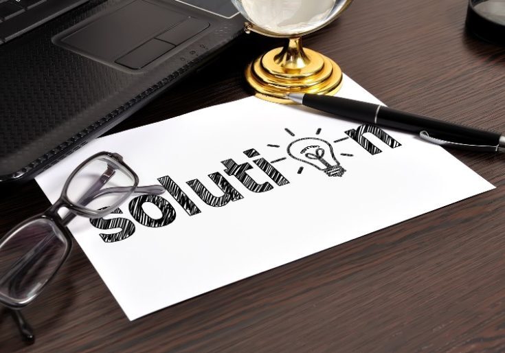 Hand-drawn 'solution' text with lightbulb sketch on white paper, surrounded by laptop, glasses, and gold desk clock