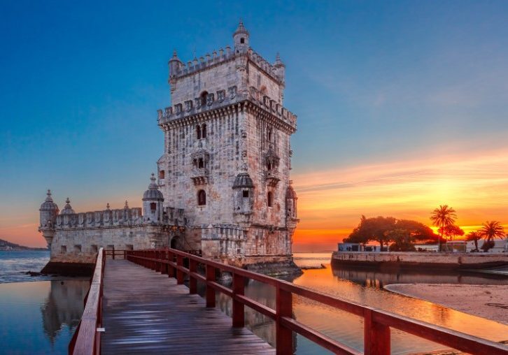 Historic Belém Tower in Lisbon, Portugal stands against a vibrant orange sunset sky, reflected in calm waters.