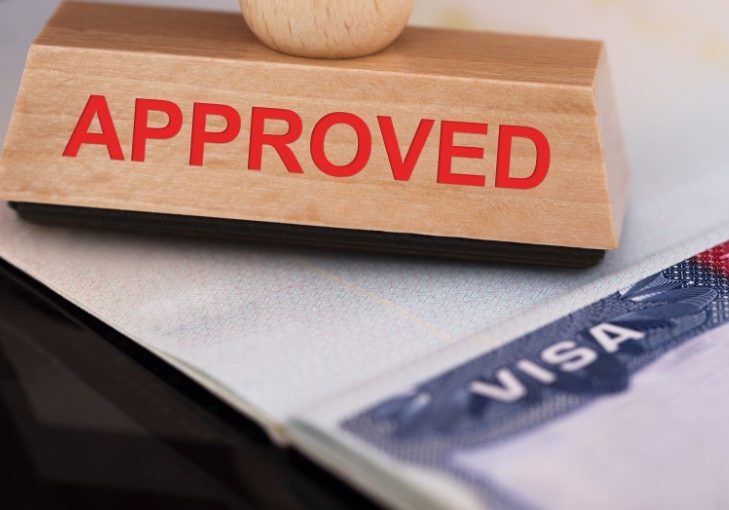 Wooden "APPROVED" rubber stamp positioned above a partial view of a visa document